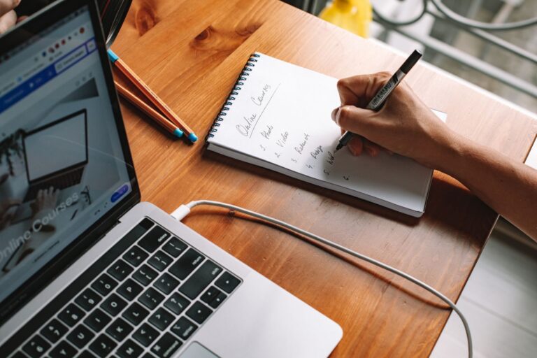 Person writing notes beside a laptop while researching