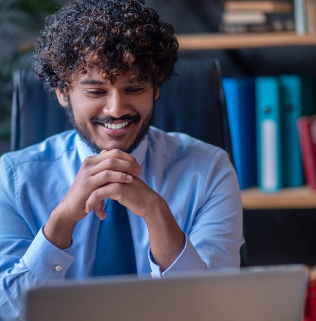 Optimism. Bearded young man with dark curly hair holding his hands near beard sitting in office at table smiling looking at laptop screen